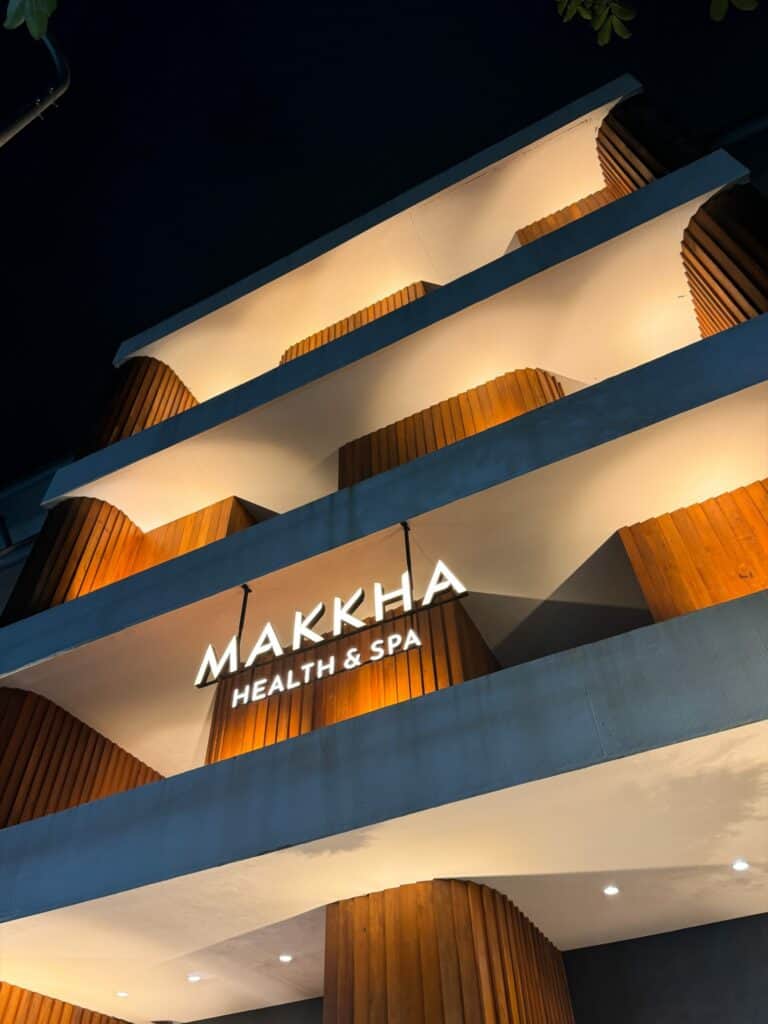 MAKKHA 健康水療中心的標誌在夜晚照亮了現代建築的外觀，該建築擁有弧形混凝土陽台和溫暖的木質鑲板。.