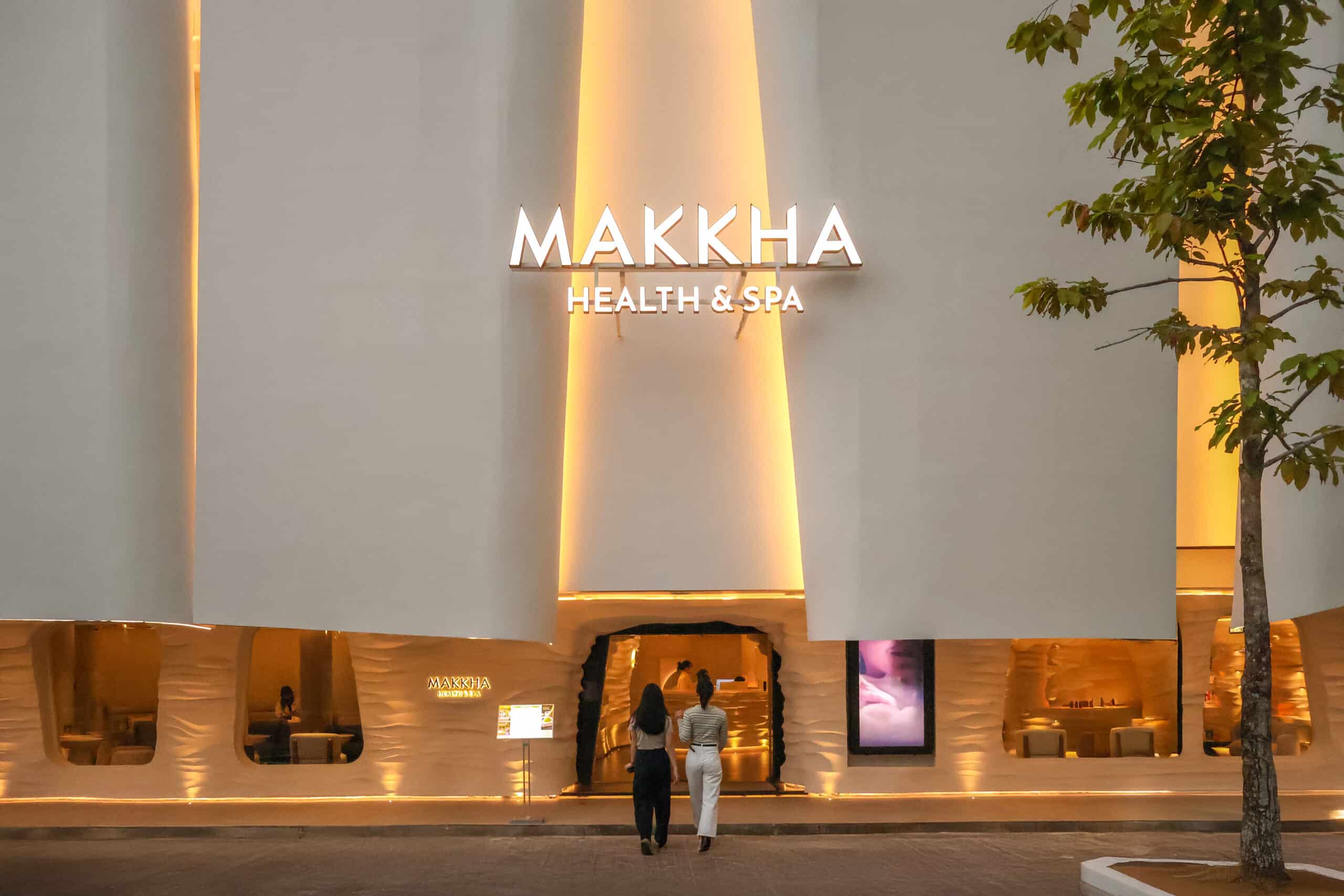 MAKKHA 健康水療中心的外觀在燈光的映襯下顯得格外醒目，現代白色外牆，溫暖的金色裝飾燈光，以及兩名女性進入水療中心的畫面。.