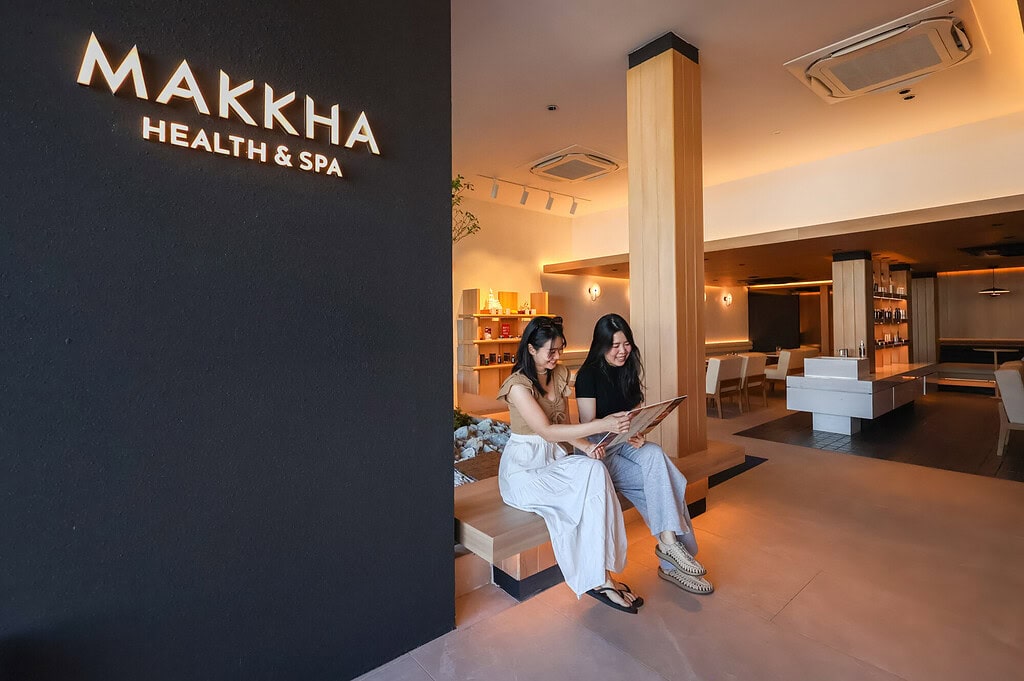 Relaxing spa lounge at Makkha Health & Spa in Chiangmai with two women enjoying a massage menu, modern interior design, warm lighting, and a welcoming atmosphere.