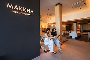 Relaxing spa lounge at Makkha Health & Spa in Chiangmai with two women enjoying a massage menu, modern interior design, warm lighting, and a welcoming atmosphere.