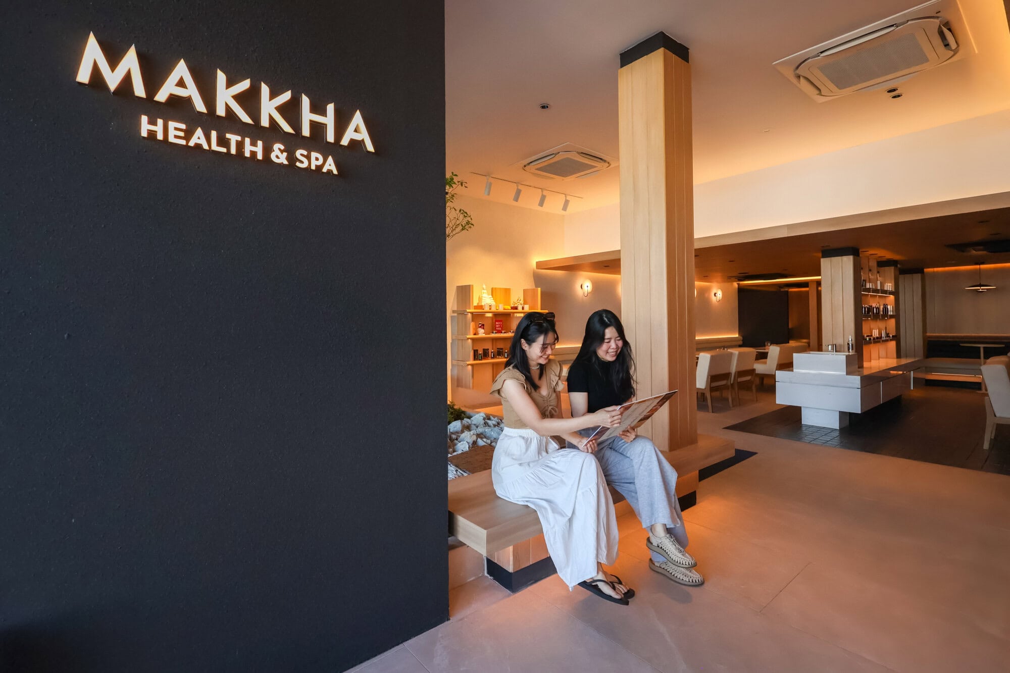 Relaxing spa lounge at Makkha Health & Spa in Chiangmai with two women enjoying a massage menu, modern interior design, warm lighting, and a welcoming atmosphere.