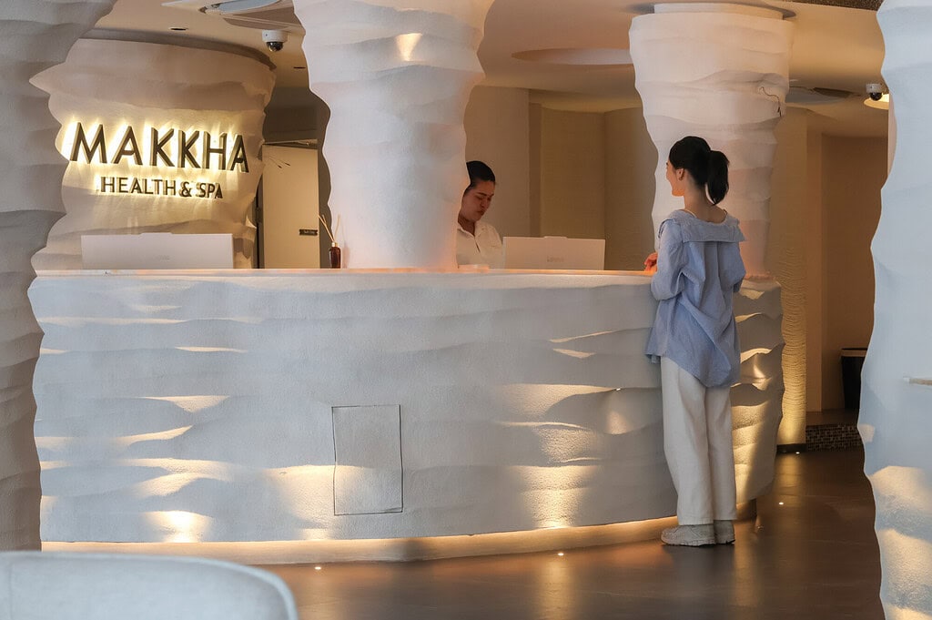 Relaxing spa reception area at Makkha Spa in Chiangmai with a friendly staff member assisting a guest, featuring modern design elements and warm lighting for a calming atmosphere.