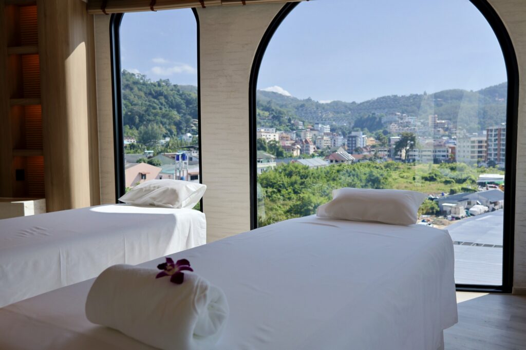 Relaxing massage room with large panoramic windows overlooking Chiangmai's scenic landscape and cityscape, featuring two massage beds with neatly arranged towels and pillows, creating a tranquil spa e.