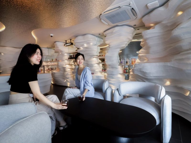 Relaxing spa lounge in Chiangmai with modern white textured walls, comfortable seating, and two women enjoying drinks, creating a serene environment at Makkha Spa.